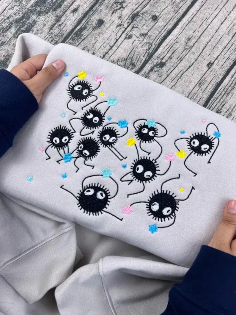 May include: A light gray sweatshirt featuring an embroidered design of several black soot sprites with white eyes and colorful accents. The design includes yellow, pink, and blue details. The sweatshirt is held by a person.