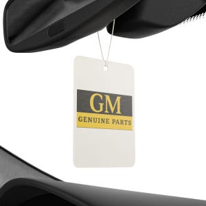Puede incluir: Una etiqueta rectangular blanca con el texto "GM" en un rect&aacute;ngulo gris oscuro y "GENUINE PARTS" en amarillo. La etiqueta cuelga de una pieza negra de un coche, probablemente un espejo retrovisor.