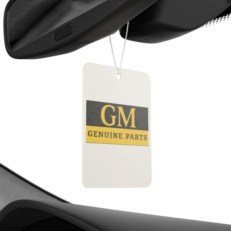 Puede incluir: Una etiqueta rectangular blanca con el texto "GM" en un rect&aacute;ngulo gris oscuro y "GENUINE PARTS" en amarillo. La etiqueta cuelga de una pieza negra de un coche, probablemente un espejo retrovisor.