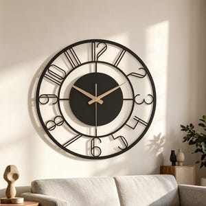 May include: A large circular wall clock featuring a black metal frame with open-work numerals from one to twelve. The center consists of a solid black circular dial with thin, gold-colored hour and minute hands, creating a stark contrast against the wall.