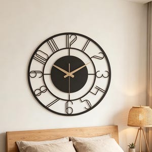 May include: A large circular wall clock featuring a black metal frame with open-work numerals from one to twelve. The center consists of a solid black circular dial with two thin, gold-colored hands indicating the time against the dark background.