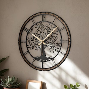 Könnte beinhalten: Eine runde Wanduhr aus schwarzem Metall mit einem Lebensbaum-Design. Die Uhr hat römische Ziffern und goldfarbene Zeiger. Die Uhr hat einen Durchmesser von etwa 61 cm. Sie ist an einer hellgrauen Wand befestigt.