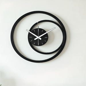 May include: A modern black wall clock with a unique spiral design. The clock features a black face with white hands and a second hand. The clock's outer frame is a large, black, circular shape, with a smaller, inner circle. The clock is mounted on a white wall.