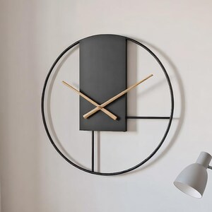 May include: A modern, minimalist wall clock with a black metal frame and a dark gray rectangular face. The clock hands are made of light-colored wood. The clock is mounted on a white wall. A gray desk lamp and a closed book are on a white table.