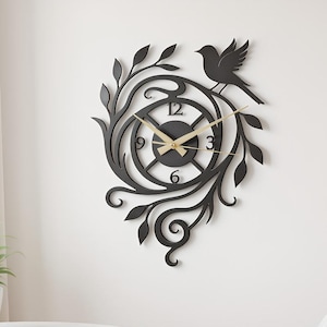 Könnte beinhalten: Schwarze Wanduhr mit Vogel- und Blumendesign. Die Uhr hat ein kreisförmiges Zifferblatt mit goldenen Zeigern und Zahlen. Die Uhr ist von dekorativen Blättern und einer Vogelsilhouette umgeben.