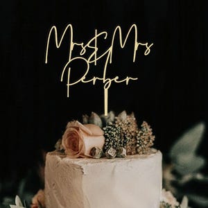May include: A gold-colored cake topper with the words "Mr & Mrs Perger" in a cursive font. The topper is placed on a tiered white cake decorated with flowers and greenery. The background is a dark, blurred setting.