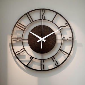 May include: A round, open-faced wall clock with a dark brown metal frame and a textured, dark brown center. The clock features white hands and Roman numeral hour markers. The clock is mounted on a light-colored wall.