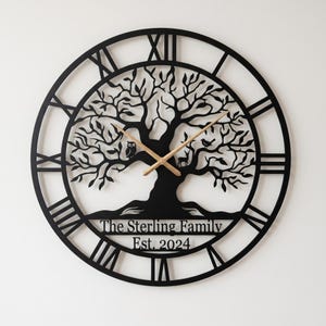 Könnte beinhalten: Schwarze Metall-Wanduhr mit einem Lebensbaum-Design. Die Uhr hat römische Ziffern und hölzerne Zeiger. Der Text "The Sterling Family Est. 2024" ist am unteren Rand der Uhr zu sehen.