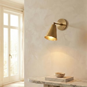 May include: A brass wall light with a conical shade and a circular backplate. The light is mounted on a light-coloured wall. The light is on, illuminating the area below. A marble table with a book and a small bowl is in the foreground.