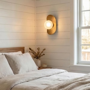 Puede incluir: Aplique de pared de color latón con una luz de globo esmerilado. El aplique tiene una forma curva y orgánica y está montado en una pared blanca con tablones. El dormitorio incluye una cama con ropa de cama blanca y una ventana.