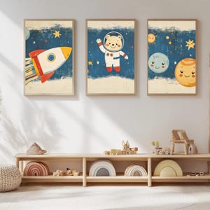 Könnte beinhalten: Drei gerahmte Kunstdrucke mit Weltraum-Motiven. Einer zeigt eine Rakete, ein anderer einen Astronauten-Bären und der dritte lächelnde Planeten. Das Kunstwerk hat einen Vintage-Look mit einer blauen und beigen Farbgebung.