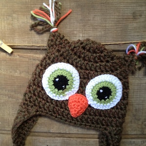May include: Brown crocheted owl hat with green and white eyes and an orange beak. The hat has long ear flaps and a brown and white yarn tassel.