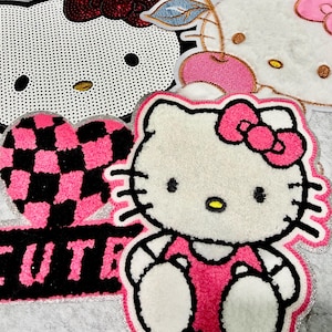 May include: Close-up of several decorative patches featuring the Hello Kitty character. The patches are in various designs, including a heart with a pink and black checkered pattern, and the word "CUTE". The patches are white, pink, and black.