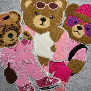 May include: Embroidered patches featuring brown teddy bears wearing sunglasses and pink outfits. One patch shows a teddy bear wearing a pink tracksuit with the text "HEAKILSW" and "LOVE LOVE LOVE". Other patches show teddy bears wearing pink shorts and hats.