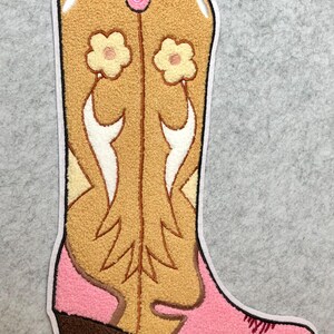 May include: A decorative patch in the shape of a cowboy boot. The boot features a tan body with white and yellow accents, a pink top and foot, and a brown heel. Two small flowers are on the boot.