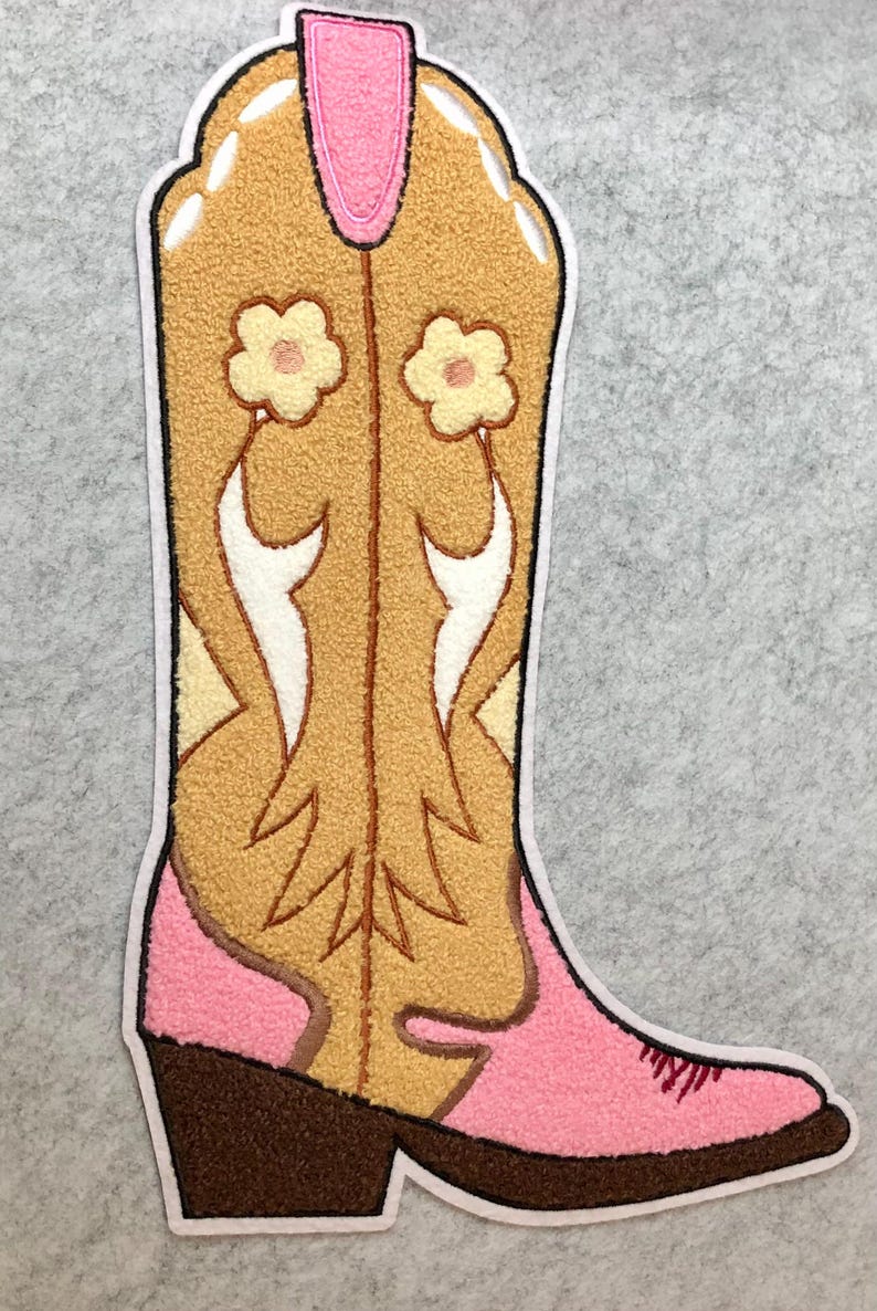 May include: A decorative patch in the shape of a cowboy boot. The boot features a tan body with white and yellow accents, a pink top and foot, and a brown heel. Two small flowers are on the boot.