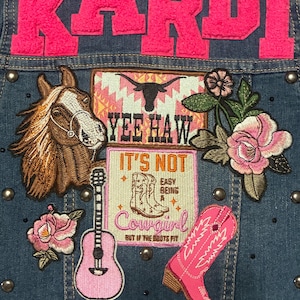 Könnte beinhalten: Jeansjacke mit rosa gestickten Aufnähern. Mit dem Namen "Kardi" in großen rosa Buchstaben. Aufnäher sind ein Pferd, eine Gitarre, ein Cowboy-Stiefel und der Schriftzug "Yee Haw". Verziert mit silbernen Nieten.