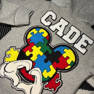 Pode incluir: Um moletom cinza com um grande design colorido de peças de quebra-cabeça em forma de rato. A palavra "CADE" é bordada em letras brancas em bloco acima do design. O moletom tem capuz e mangas compridas.