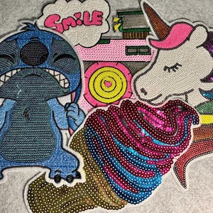 Può includere: Una collezione di toppe di paillettes colorate. Le toppe includono un personaggio dei cartoni animati blu, una macchina fotografica rosa, un unicorno con un corno rosa, un cupcake e un fumetto con la parola "SMILE".