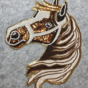 May include: A horse head applique with a gold sequin outline. The horse is made of white, brown, and gold sequins. The horse's mane and face are detailed with various sequin colors and patterns. The applique is on a gray background.