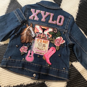 Puede incluir: Chaqueta vaquera azul con parches y tachuelas. La parte trasera de la chaqueta tiene el nombre "XYLO" en letras rosas. Los parches incluyen un caballo, una guitarra, una bota de vaquero rosa y las frases "Yee Haw" y "It's Not Easy Being a Cowgirl".