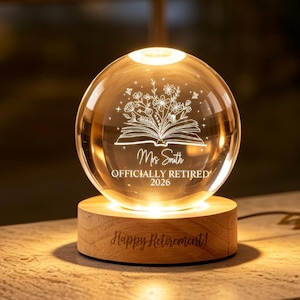Puede incluir: Una luz nocturna de esfera de cristal transparente sobre una base redonda de madera. La esfera presenta un diseño grabado de un libro abierto con flores y el texto "Ms Smith OFFICIALLY RETIRED 2026". La base de madera tiene grabado "Happy Retirement!".