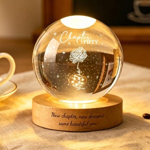Puede incluir: Una esfera de cristal transparente con un diseño floral y las palabras "Chapter Fifty" y "New chapter, new dreams, same beautiful you" grabadas. La esfera se asienta sobre una base de madera, iluminada por una luz cálida, rodeada de granos de café y una taza de café.