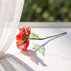 Könnte beinhalten: Eine handgefertigte Glasskulptur in Form einer Blume mit leuchtend roten Blütenblättern und einem strukturierten grünen Zentrum, befestigt an einem dünnen, dunkelgrünen Stiel mit zwei durchscheinenden grünen Blättern. Das Glas wirft einen Schatten.
