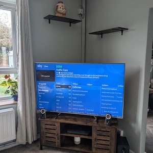 May include: A wooden TV stand with a dark finish, featuring a large flat-screen TV displaying a TV guide. Two small speakers flank the TV. A small black subwoofer sits on the floor. A decorative shelf holds a small figurine.