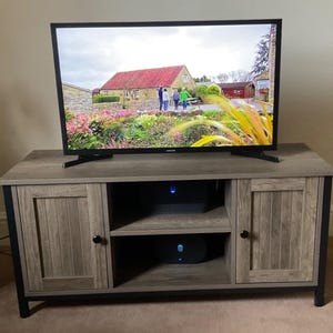 Puede incluir: Un mueble de TV gris y negro con dos puertas de armario y estanterías abiertas. El mueble de TV tiene un acabado de vetas de madera y detalles metálicos negros. Un televisor Samsung muestra una colorida escena al aire libre.