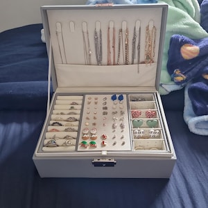 Op de afbeelding: Open grijze sieradendoos met meerdere compartimenten. Het bovenste gedeelte bevat kettingen, terwijl de onderste gedeelten ringen, oorbellen en andere kleine sieraden bevatten. De doos is open en toont diverse sieraden.