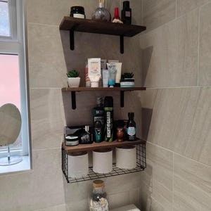 Può includere: Mensole da bagno con vari articoli da toeletta e oggetti decorativi. Le mensole sono in legno scuro e sostenute da staffe in metallo nero. Gli oggetti includono lozioni, piante e un barattolo di vetro pieno di conchiglie.