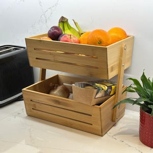 May include: A two-tiered bamboo fruit basket containing fresh produce. The top level has oranges, bananas, an apple, and a plum. The bottom level holds an onion and a packaged food item. A black toaster is in the background.