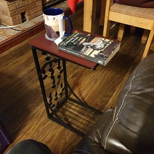 Op de afbeelding: Een kleine, donkerrode bijzettafel met een zwart metalen frame. Een boek en een mok staan op de tafel. De tafel heeft een decoratief bladerontwerp en staat naast een bruine leren bank.