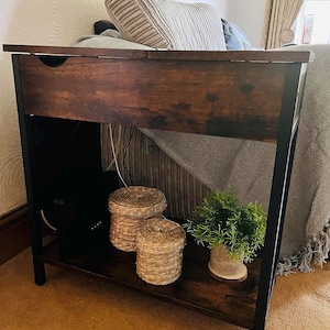 May include: A dark brown wooden side table with a black metal frame. The table has a drawer and a lower shelf. Two woven baskets and a small potted plant sit on the shelf. The table is 30 inches tall.