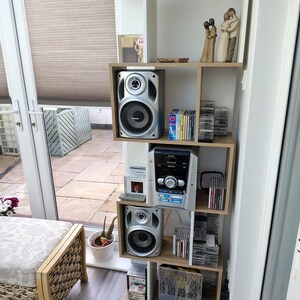 Może przedstawiać: Drewniany regał z białym i jasnobrązowym wykończeniem, mieszczący wieżę stereo, głośniki, płyty CD i elementy dekoracyjne. Głośniki są srebrno-czarne. Na pierwszym planie ławka z wzorzystą poduszką.