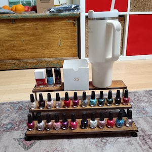 May include: A wooden tiered nail polish display stand with various nail polish bottles in different colours. A large white tumbler with a handle is on the right. A white box with the number 25 is also present.