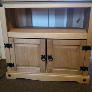 May include: A light-colored wooden media console with two cabinet doors and an open shelf. The cabinet doors have black metal ring pulls and hinges. The console has a curved base and black decorative accents.