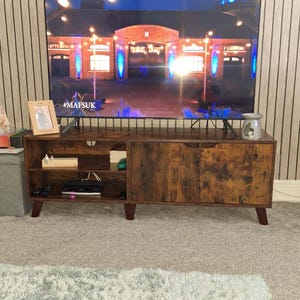 Puede incluir: Un mueble de TV marrón oscuro con una puerta corredera y estantes abiertos. El mueble tiene un acabado envejecido y está sobre patas cortas y anguladas. Un televisor grande muestra una escena brillantemente iluminada. Una foto enmarcada y un difusor encima.