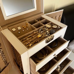 May include: A white jewelry box with multiple compartments and drawers, filled with various pieces of jewelry. The top section features a mirror and is lined with a tan fabric. The drawers are open, revealing necklaces, rings, earrings, and bracelets in gold and silver tones.