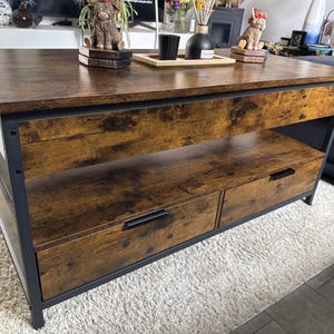 Puede incluir: Mesa de centro rectangular de color marrón oscuro con efecto veteado. Cuenta con un estante inferior y dos cajones con tiradores negros. La mesa tiene una estructura de metal negro y está sobre una alfombra clara. La mesa mide unos 120 cm de largo.