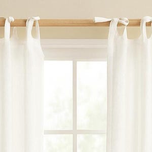 May include: Two white linen curtains suspended from a natural wood rod. The curtains are attached to the rod with white fabric loops. A window with white trim is visible between the curtains.
