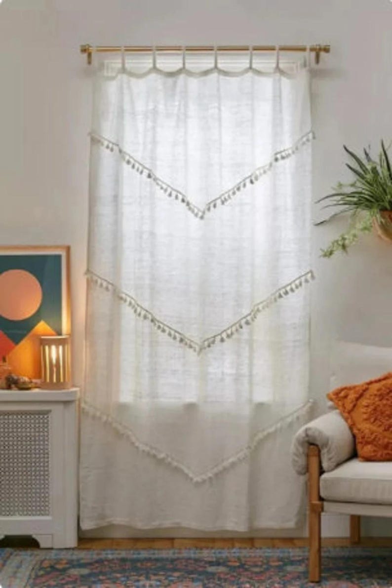 May include: White linen curtain with a scalloped top and chevron-shaped tassel trim. The curtain hangs in front of a window, with a gold-colored curtain rod. The room has a bohemian style, with a rug and a chair.