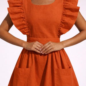 May include: An orange dress with a square neckline and frilled sleeves. The dress has a fitted bodice, a wide waistband, and a flared skirt with two large pockets. The fabric looks like linen or a similar natural material.