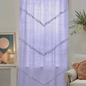 May include: A light purple curtain with a rod at the top. The curtain has a series of inverted V-shaped designs with tassels. The curtain is hanging in front of a window. The curtain is made of a light, airy fabric.