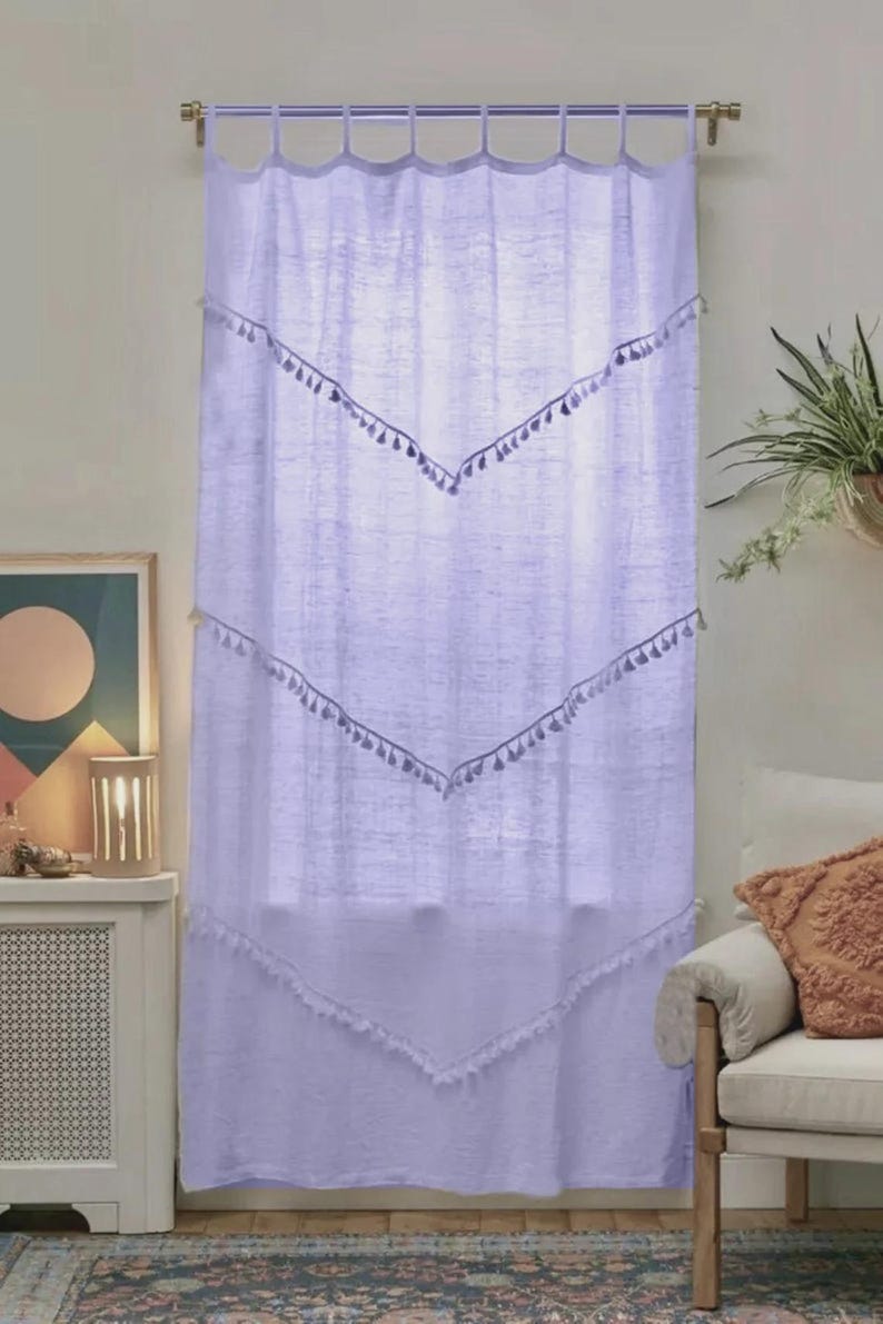May include: A light purple curtain with a rod at the top. The curtain has a series of inverted V-shaped designs with tassels. The curtain is hanging in front of a window. The curtain is made of a light, airy fabric.