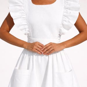 May include: A white dress with a square neckline and ruffled sleeves. The dress has a fitted bodice and a flared skirt with two large pockets. The dress is made of a lightweight fabric, ideal for summer.