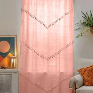 May include: A light peach-colored curtain with a rod at the top. The curtain has a series of chevron-shaped designs with tassels. The curtain is hanging in front of a window. The curtain is made of a light, airy fabric.