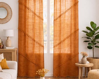 May include: Two burnt orange linen curtains with diagonal tassel trim hang in front of a window. The curtains are hung on a gold rod. The room has a neutral color palette with a round wooden coffee table.