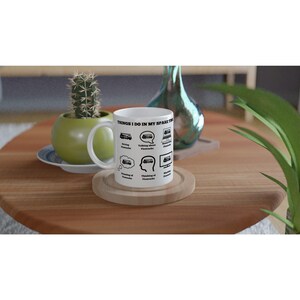 Peut inclure: Tasse en céramique blanche avec le texte "THINGS I DO IN MY SPARE TIME" et des illustrations de bus. La tasse est posée sur un dessous de verre en bois. Un petit cactus dans un pot vert et un vase décoratif en verre sont en arrière-plan.